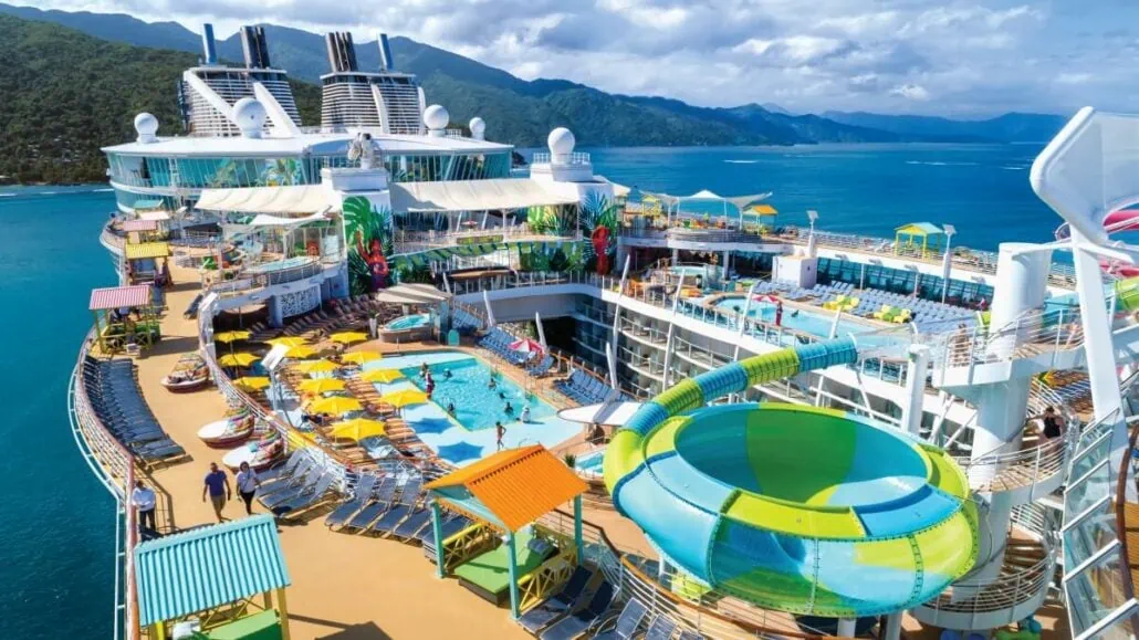 Aerial view of a cruise ship's deck featuring pools, water slides, sunbathing areas with lounge chairs, and a scenic ocean background with hills—a true $100M glow-up courtesy of Royal Caribbean.
