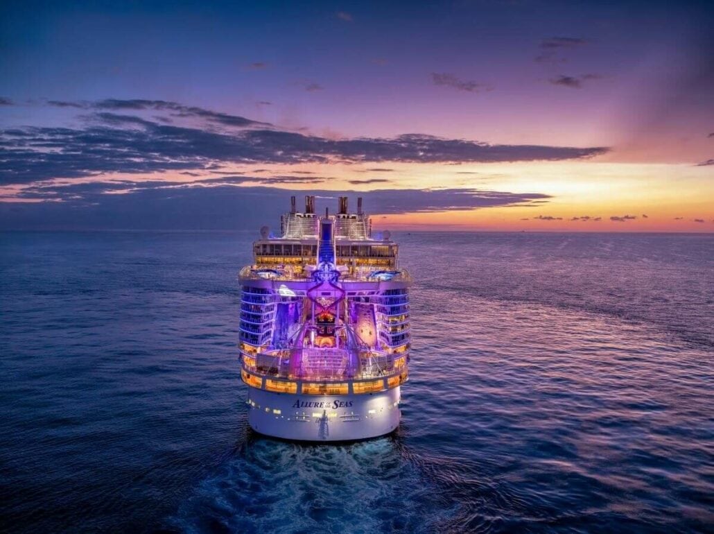 A Royal Caribbean cruise ship, boasting a $100M Glow-Up, sails on a calm ocean at sunset, its vibrant purple and blue lights illuminating the scene against a backdrop of clouds and clear sky.