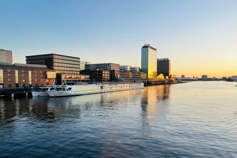 A river scene at sunset in Amsterdam with buildings along the waterfront and a docked riverboat, reflecting on the calm water. Contingency plans ensure smooth operations as cruise protests occasionally bring attention to the bustling harbor.