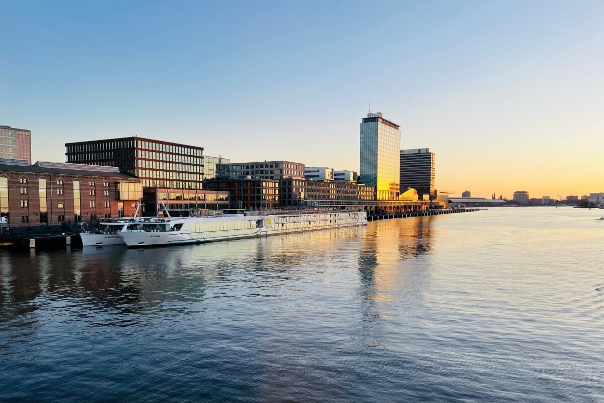 A river scene at sunset in Amsterdam with buildings along the waterfront and a docked riverboat, reflecting on the calm water. Contingency plans ensure smooth operations as cruise protests occasionally bring attention to the bustling harbor.