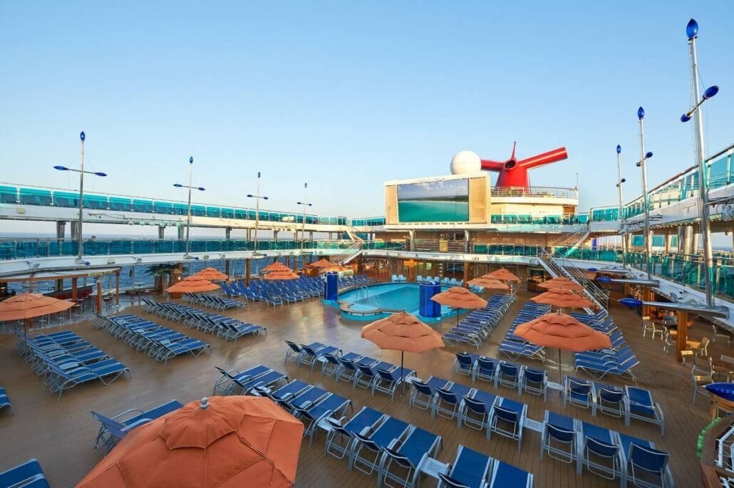 A cruise ship's outdoor deck features a swimming pool, numerous lounge chairs, and orange umbrellas, with a large screen in the background. Amidst the leisure, news of a missing Carnival crew member had prompted discussions about maritime search and rescue operations.