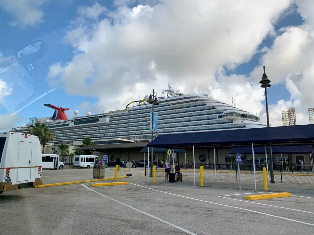 A large white cruise ship is docked at the Galveston port in Texas. Nearby, several buses and vehicles from Best Cruise Parking are stationed. The sky is partly cloudy. Yellow bollards and a covered walkway are visible in the foreground, where a person with luggage stands near a pole.