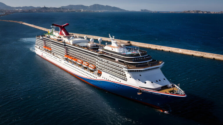 A large cruise ship sails near a coastal pier with mountainous terrain visible in the background and a clear blue sky overhead, part of the exciting 2026 itineraries offered by Carnival.