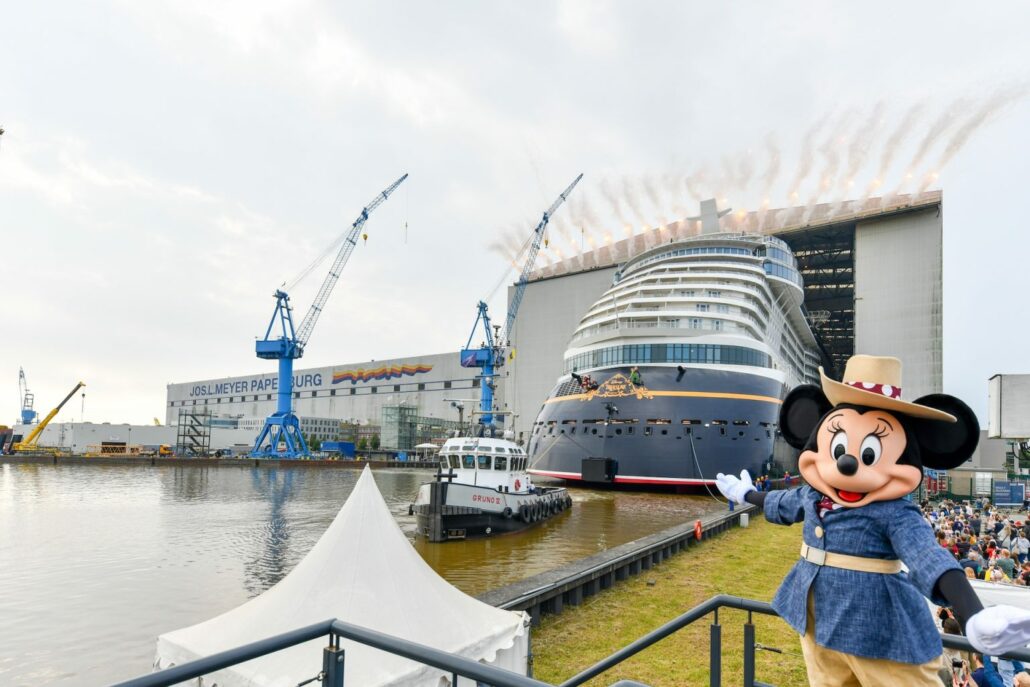 A large cruise ship, the Disney Treasure, is being launched at a bustling shipyard, with a person in a cartoon-character costume waving from the foreground. Cranes and industrial buildings are visible in the background as the majestic vessel floats out for its maiden voyage.