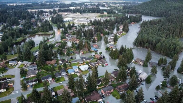 Aerial view of a residential area in Juneau heavily flooded with water encroaching on houses and streets, surrounded by dense forest. In this Norwegian landscape, a few elevated areas remain dry, highlighting the urgent need for flood-relief efforts.