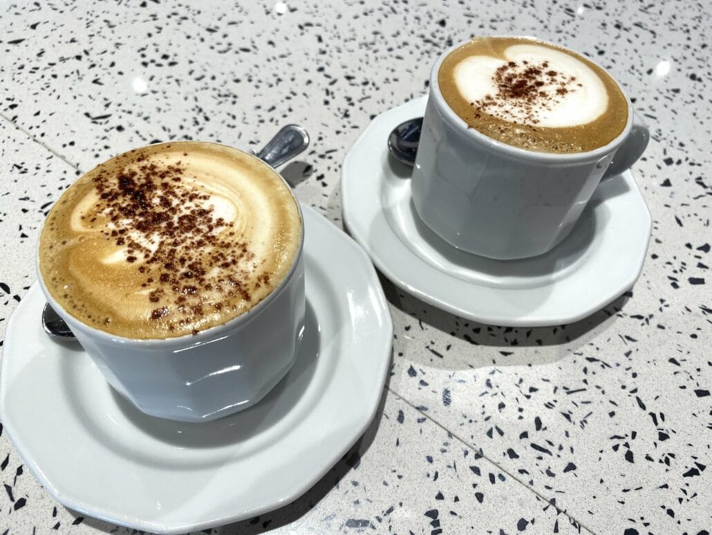 Two cappuccinos in white cups on saucers sit on a speckled countertop, reminiscent of mornings aboard MSC Cruise with its drink packages. Each coffee boasts frothy milk crowned with cocoa powder, while spoons rest elegantly beside them.