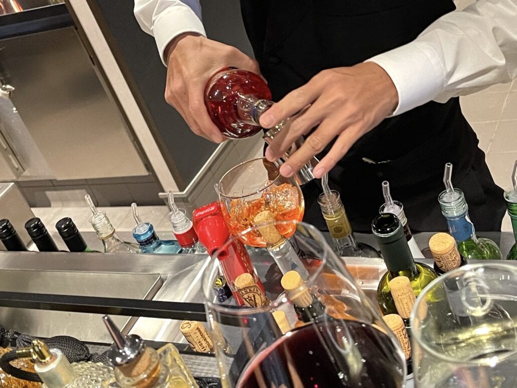 A bartender, clad in a white shirt and black vest, pours a red liquid into a glass amid an array of liquor bottles, syrups, and corks. This vibrant scene could easily be part of the dynamic offerings found in MSC Cruise Drink Packages. The setting exudes the allure of a classic bar experience.