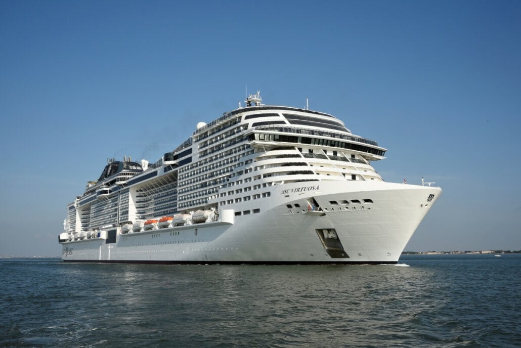 A large white cruise ship sails on the ocean, showcasing multiple decks and lifeboats on a clear, sunny day. Amidst the luxurious journey, whispers of jewelry theft add an unexpected twist to the serene adventure.