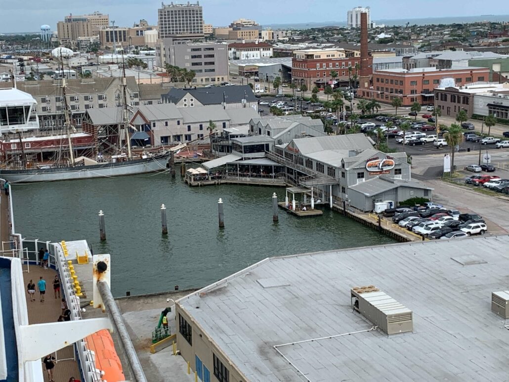 Aerial view of the coastal city of Galveston, Texas, with a bustling harbor. Boats are docked along the waterfront near cruise parking facilities, surrounded by buildings and parking lots. The diverse city skyline stands silhouetted in the background under an overcast sky.