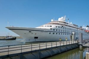 A large white cruise ship named Star Legend, part of the Windstar fleet, docked at a port, with two individuals in safety vests walking on the adjacent pier.