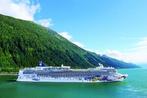 A large white Norwegian cruise ship with colorful designs on its hull sails on calm, greenish water near Juneau's mountainous landscape covered in dense greenery under a clear blue sky.