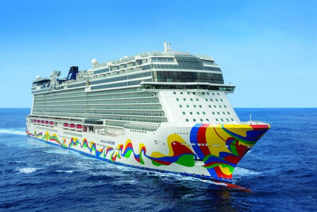 A large cruise ship, the Norwegian Encore, with colorful patterns painted on its hull, sails on a calm, blue sea under a clear sky. The vessel's recent enhancements add to its majestic presence on the serene waters.