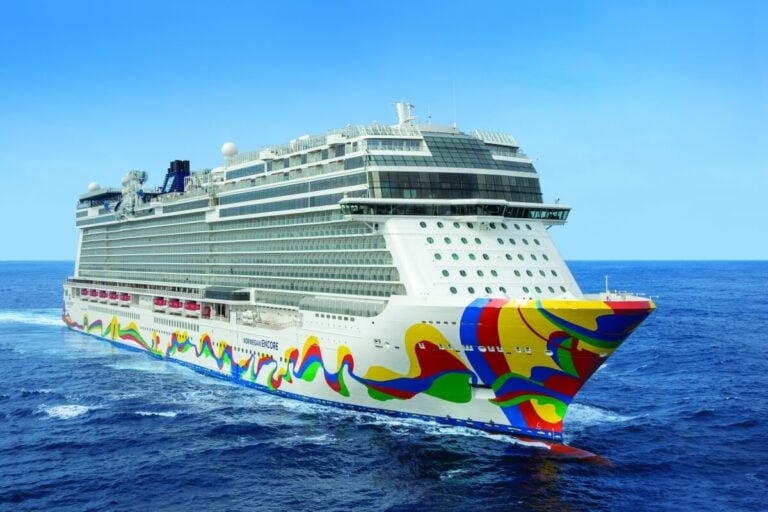 A large cruise ship, the Norwegian Encore, with colorful patterns painted on its hull, sails on a calm, blue sea under a clear sky. The vessel's recent enhancements add to its majestic presence on the serene waters.