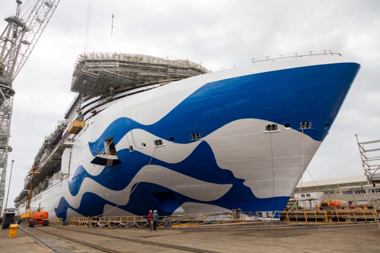 Star Princess Sphere-Class ship with a blue and white wave pattern painted on the hull is docked in a shipyard. Two workers in hard hats and safety vests stand nearby. Scaffolding and cranes are visible around the ship, marking this major milestone. The sky is overcast.