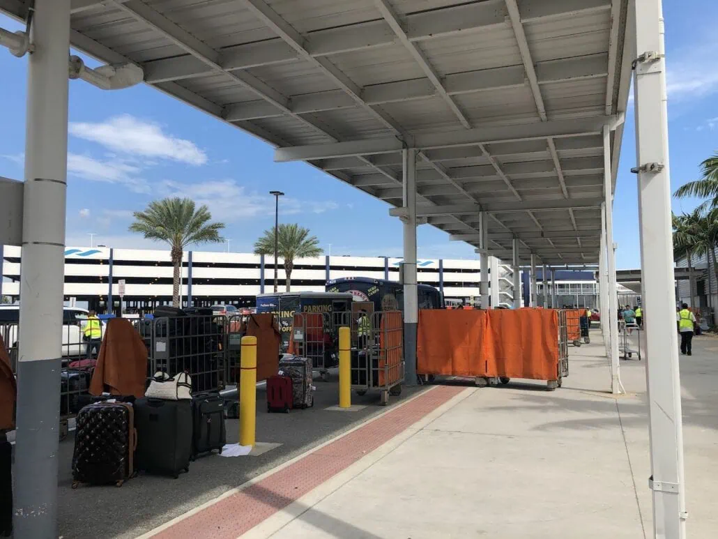 A view of an airport area with luggage carts lined up under a canopy. Several suitcases and bags are placed in and around the carts, covered by orange tarps. Palm trees are visible in the background, and a parking structure is seen across the street. A baggage handler's cart rolls by quietly.