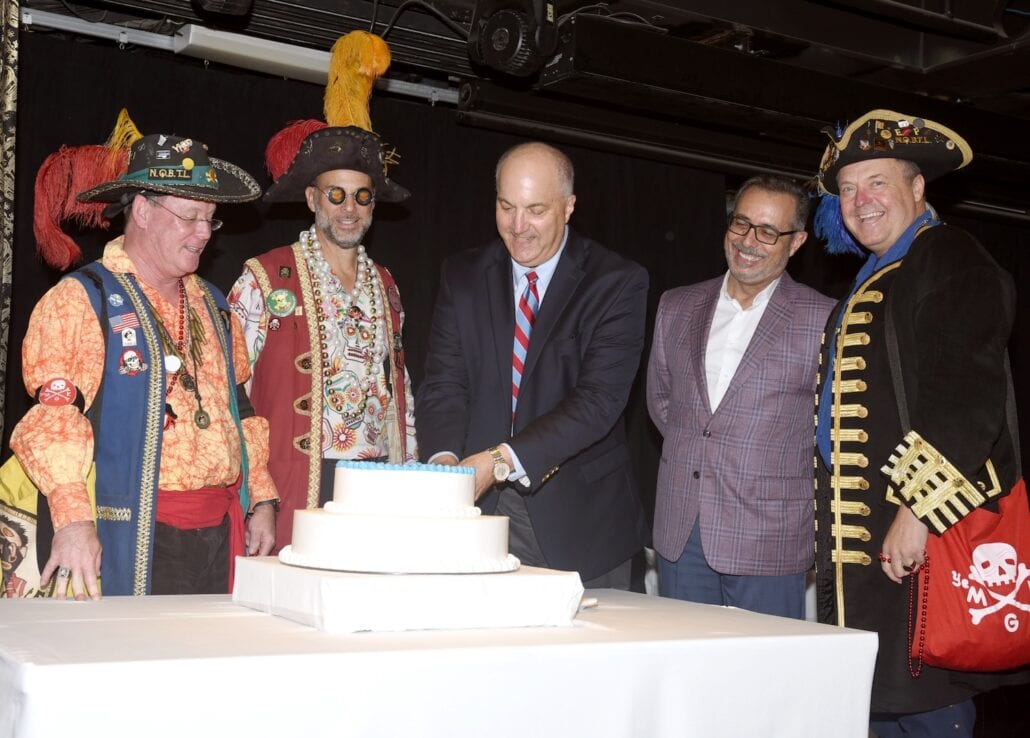 A group of men stand around a white table with a cake. One man in a suit and tie is cutting the cake, while three men in colorful pirate costumes and one man in a blazer watch, likely celebrating after the Tampa Home Port beach clean-up event indoors.