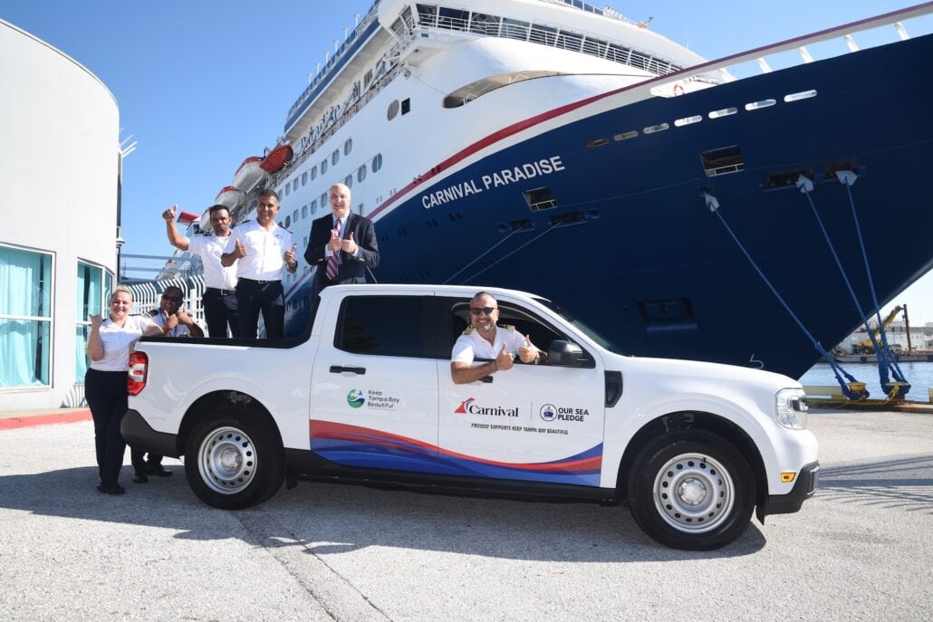 A group of five people in uniforms are posing with a white pickup truck labeled "Carnival Celebrates Tampa Home Port" and "Port of Tampa Bay" in front of a large docked cruise ship named "Carnival Paradise." One person is sitting in the driver’s seat with a thumbs-up.