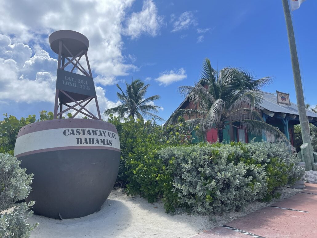 A signpost at Castaway Cay, Bahamas with latitude 26.1 and longitude 77.5 markings catches the eye. Surrounded by lush green bushes, palm trees, and a small building in the background under a partly cloudy blue sky, it's a perfect photo op during your Disney Fantasy Cruise adventure.