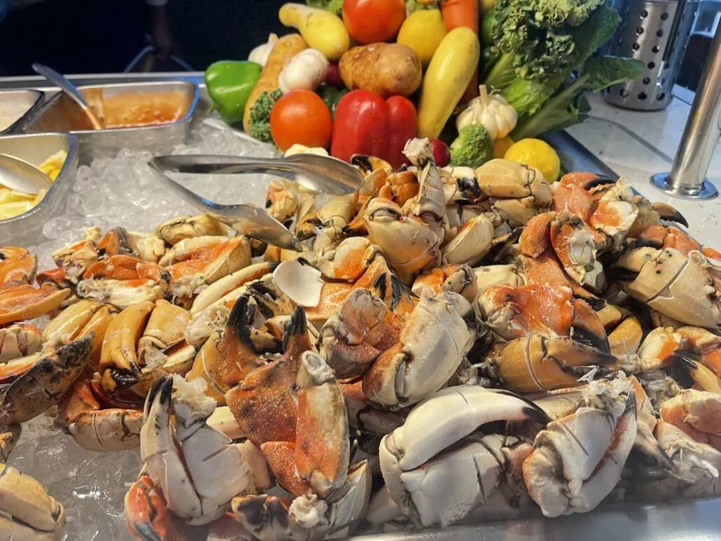 A seafood display on the Disney Fantasy Cruise showcases a pile of crab claws resting on ice. Behind the crabs, colorful vegetables like bell peppers, potatoes, tomatoes, broccoli, and garlic are artistically arranged. Metal tongs are conveniently placed on top of the crab claws for easy serving. Bon voyage!