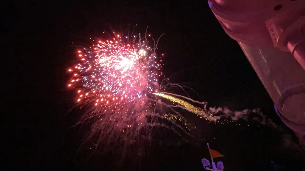 A vibrant fireworks display in the night sky with a mix of red and yellow streaks. The structure to the right has a cylindrical shape and is illuminated by the fireworks. Small bursts of light are visible near the main explosion, creating a magical scene reminiscent of Disney Fantasy Cruise tips for an unforgettable voyage.