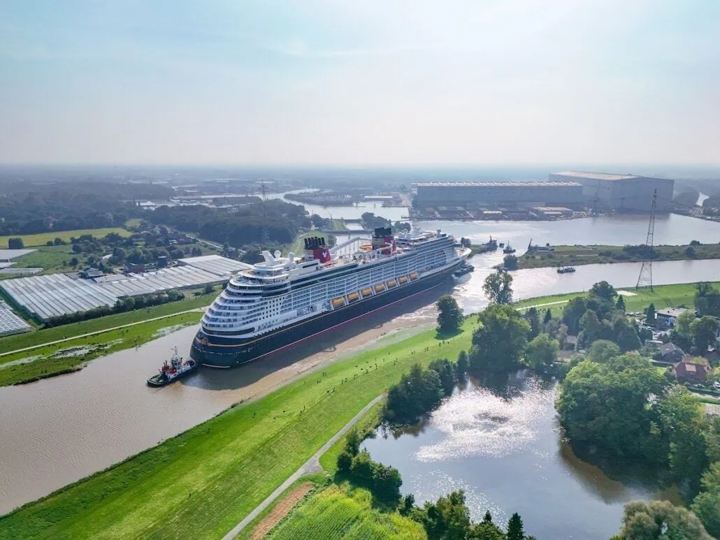 The Disney Treasure, fresh from sea trials at the shipyard, is navigating a narrow river channel near a green shoreline, with industrial buildings and farmlands in the background on a hazy day.