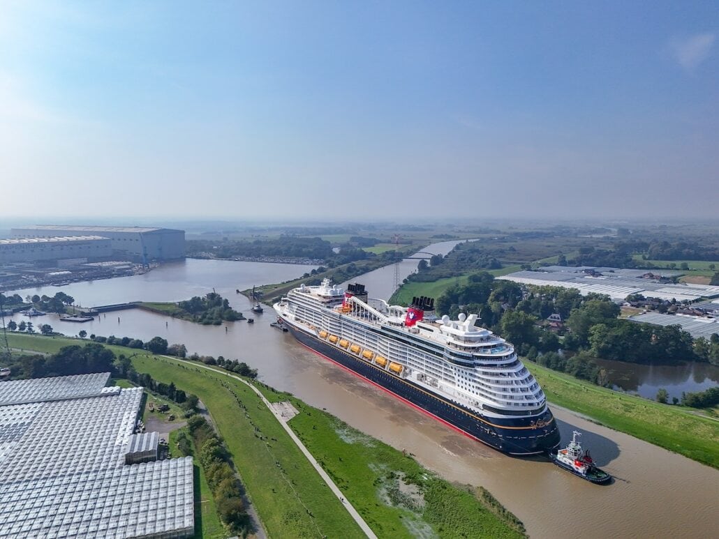 An aerial view showcases the Disney Treasure, a large cruise ship navigating through a river flanked by green fields and industrial buildings, accompanied by a small tugboat as it prepares for sea trials near the shipyard.