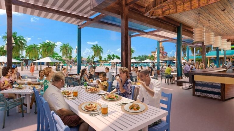 A group of people is dining at an outdoor restaurant with tropical decor. Large wooden beams support a covered area, protecting patrons from the sun. In the background, there are palm trees, umbrellas, and a sunny blue sky indicating a beachside location.