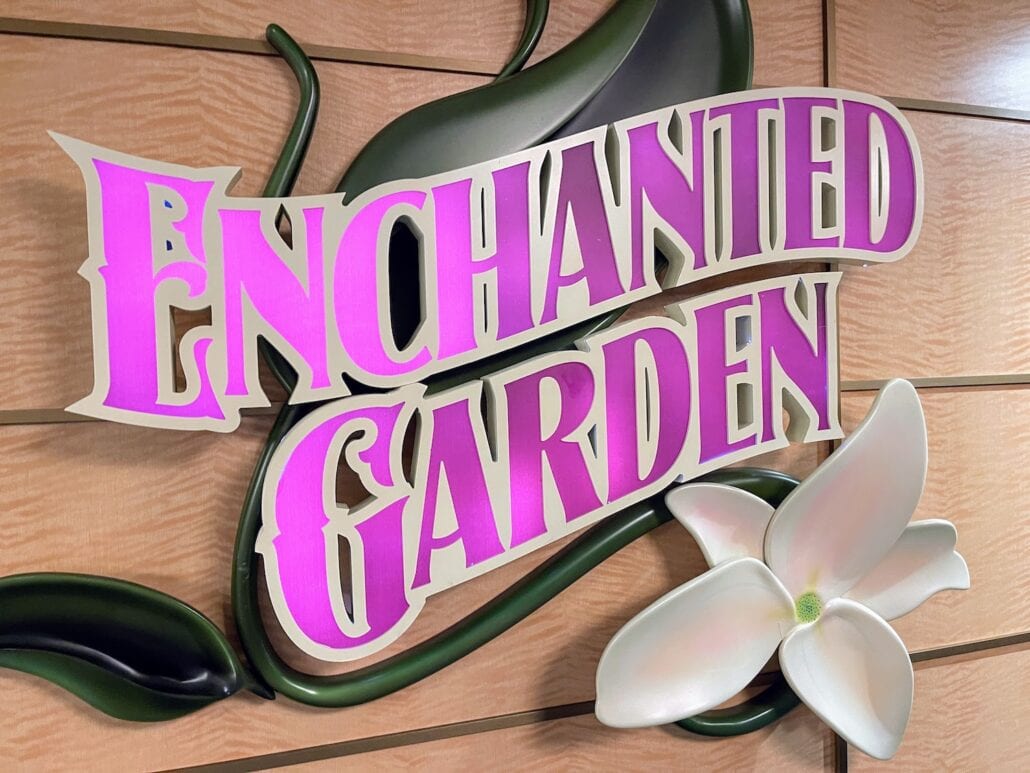 A sign with the text "Enchanted Garden" is displayed against a wooden background. The text is colored in shades of purple and pink, enhanced by a stylized green leaf and a white flower in the bottom right corner, reminiscent of magical decor you'd find on a Disney Fantasy Cruise.