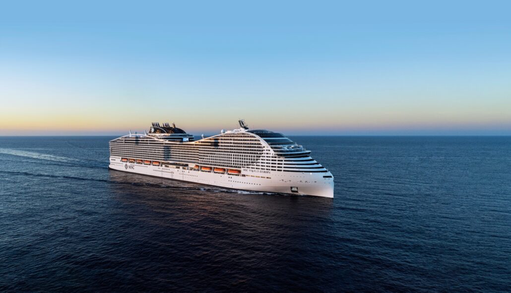A large white cruise ship, MSC World America, sails in the open ocean under a clear sky. The ship is moving away from the viewpoint, leaving a trail in the calm water. The horizon and the gradient of the sky from blue to light orange are visible in the background.