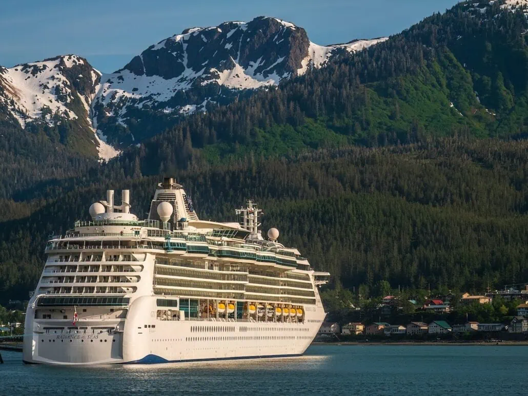 A white Royal Caribbean cruise ship equipped with Starlink internet sails in calm waters in front of snow-capped mountains and a forested hillside with scattered buildings near the Juneau cruise port.
