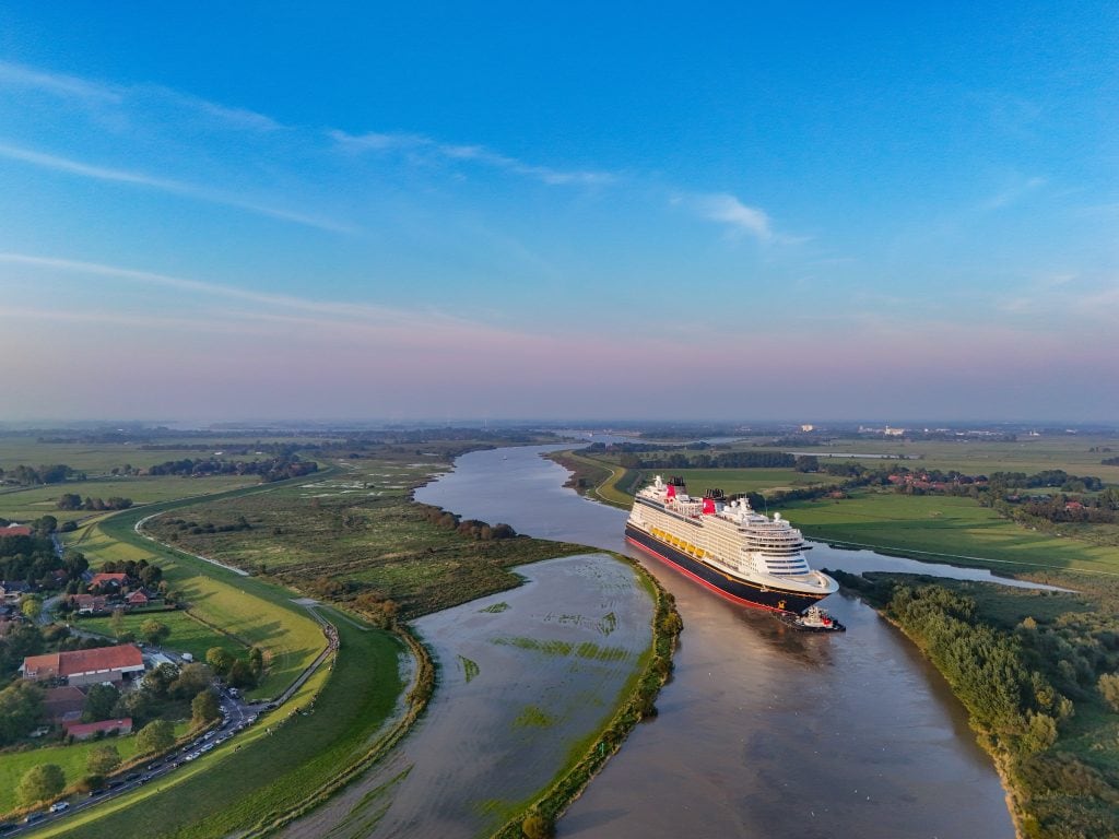 A large cruise ship, the Disney Treasure, sails along a winding river with green fields and small buildings on either side, under a clear blue sky. The vessel's sea trials were completed at a nearby shipyard just last month.