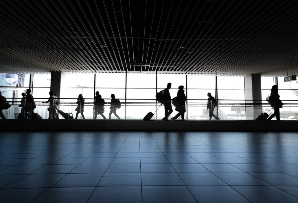 Silhouettes of families and travelers, including kids with luggage, wander through the bustling airport terminal, illuminated by large windows. The tiled floor and slatted ceiling panels create a rhythmic backdrop for bustling cruisers as they exchange travel tips.