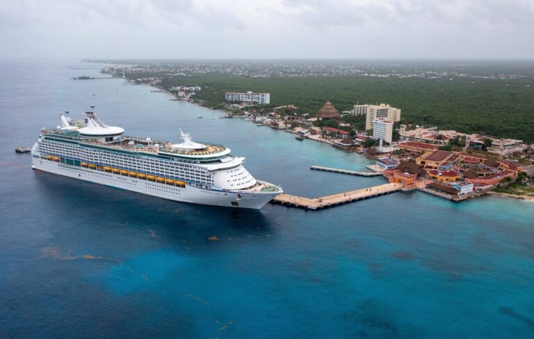 A large Royal Caribbean cruise ship docked at a pier beside the coastal town of Labadee, Haiti, with several buildings surrounded by clear blue ocean water and dense greenery in the background. This picturesque scene will be even more inviting come October 2024.