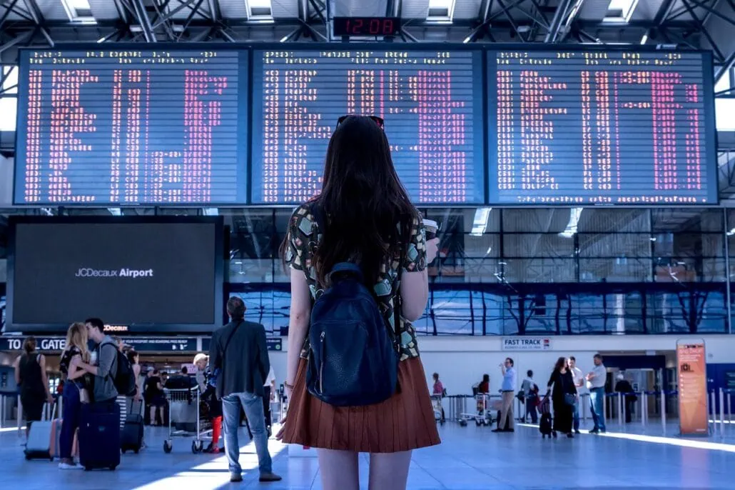 A person with long hair and a backpack stands in an airport terminal, casually observing the large flight information board. Other travelers, cruisers with kids in tow, and bustling airport facilities form the vibrant backdrop of this travel scene.