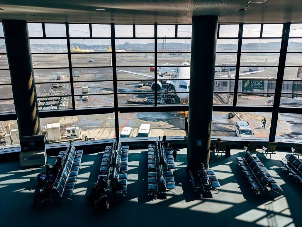 An airport terminal with large windows showing a parked airplane at the gate. Rows of empty seats are in the foreground with a few people sitting. Various airport vehicles and ground crew are visible outside. The snowy landscape in the background offers serene views for cruisers with kids awaiting their family cruise.