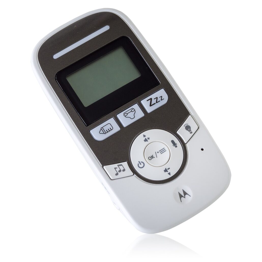 A handheld electronic device with a small rectangular screen, several buttons including a musical note, microphone, and moon with "ZZZ". Primarily white with a dark area around the screen and controls, this baby monitor features the brand logo "Motorola" at the bottom.