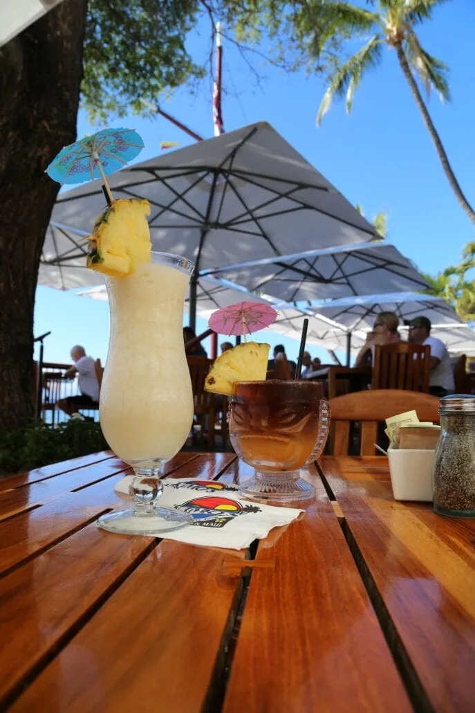 Two cocktails on a wooden table at an outdoor restaurant. The drinks are garnished with pineapple slices and small paper umbrellas. In the background, cruise passengers sit at tables under large umbrellas, with trees and a clear blue sky overhead—one of the must-do activities in Key West.