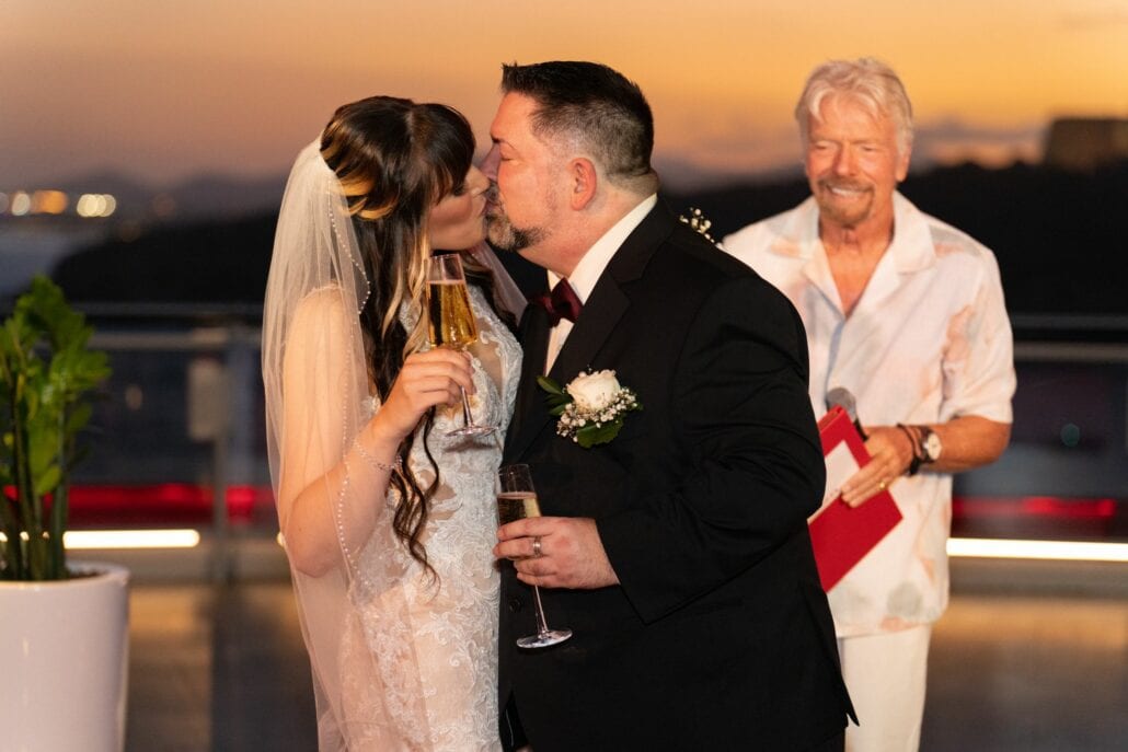 A newlywed couple shares a kiss while holding champagne glasses during their surprise wedding ceremony at sunset aboard a Virgin Voyages ship. In the background, a person in a casual shirt smiles and holds a book.
