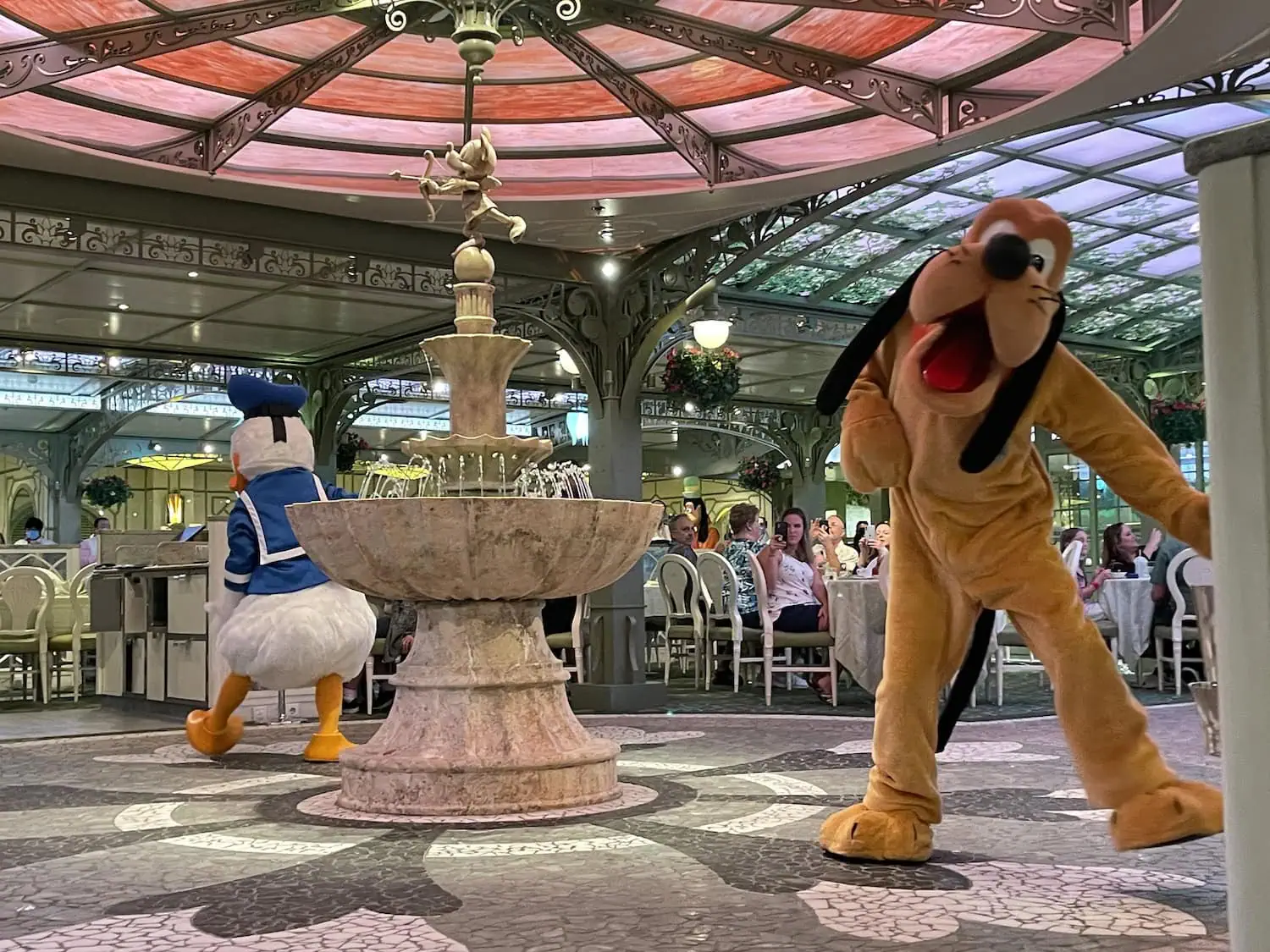 Two people in costumes of cartoon characters, one of Donald Duck and one of Pluto, perform near a central fountain in a decorated indoor space on the Disney Fantasy Cruise. The area has a glass ceiling and several tables with people dining in the background.