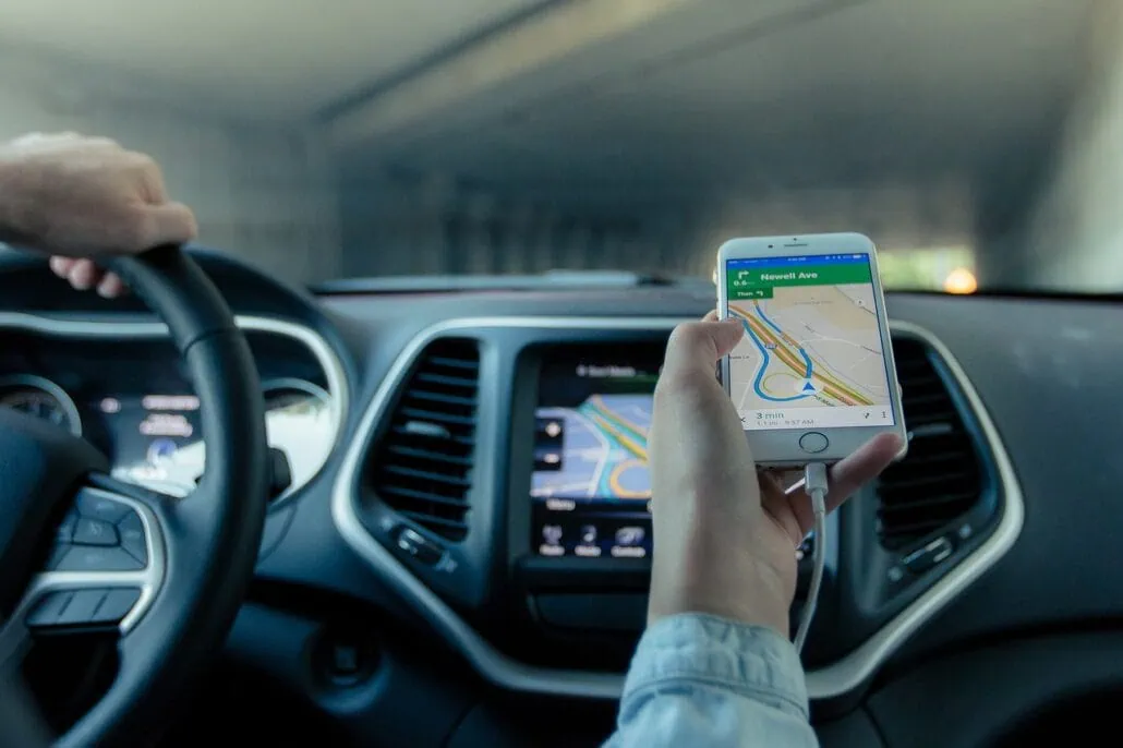 From the back seat, you watch as the driver skillfully maneuvers the car, navigating with a smartphone displaying a map towards the Port of Galveston. The dashboard reveals a built-in screen, while their steady hand remains on the steering wheel.