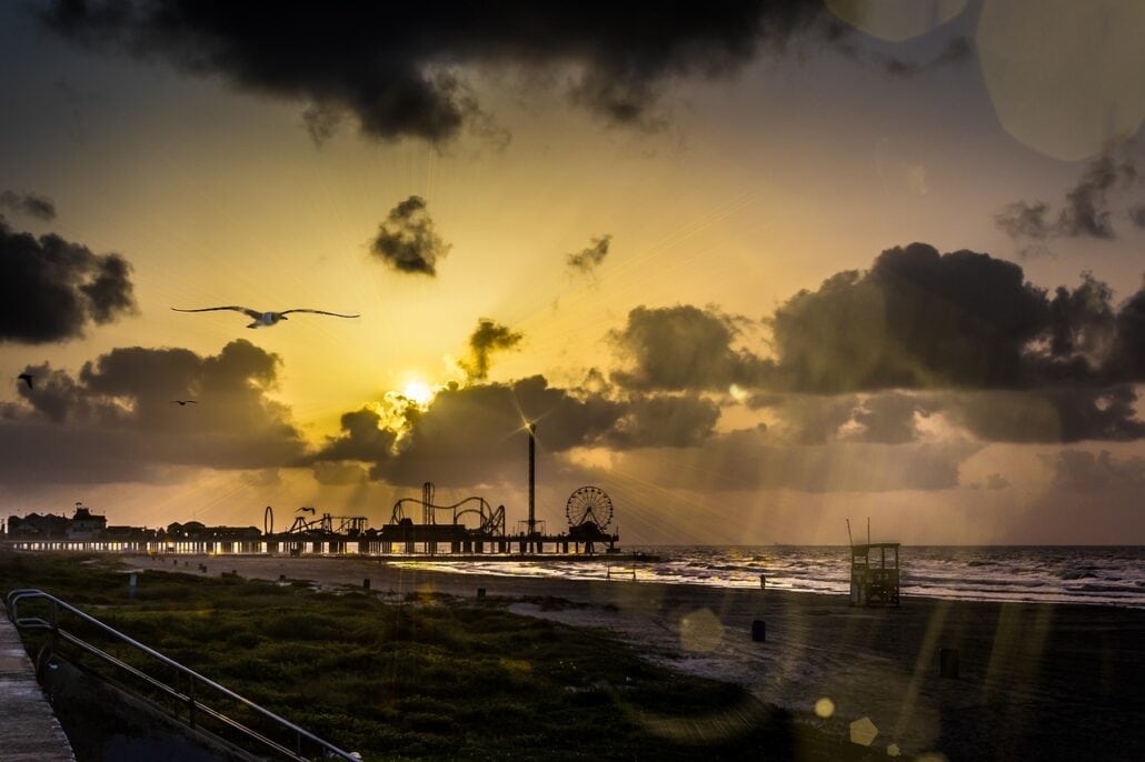 A coastal amusement park pier extends into the ocean at sunset, with various rides silhouetted against the sky. A few seagulls fly above, and the beach in the foreground shows some grassy areas and a lifeguard tower. Dark clouds partially obscure the sun—one of many things to do while visiting Galveston Island.