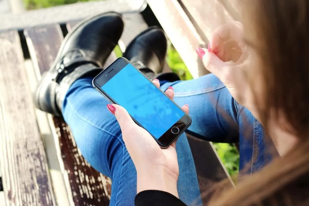 A person holding a smartphone with a blue screen sits on a wooden bench, engaged in exploring Essential Cruise Tips. They wear blue jeans and black boots, and their nails are painted red. The view from above focuses on the phone as they prepare for a smooth sailing experience.
