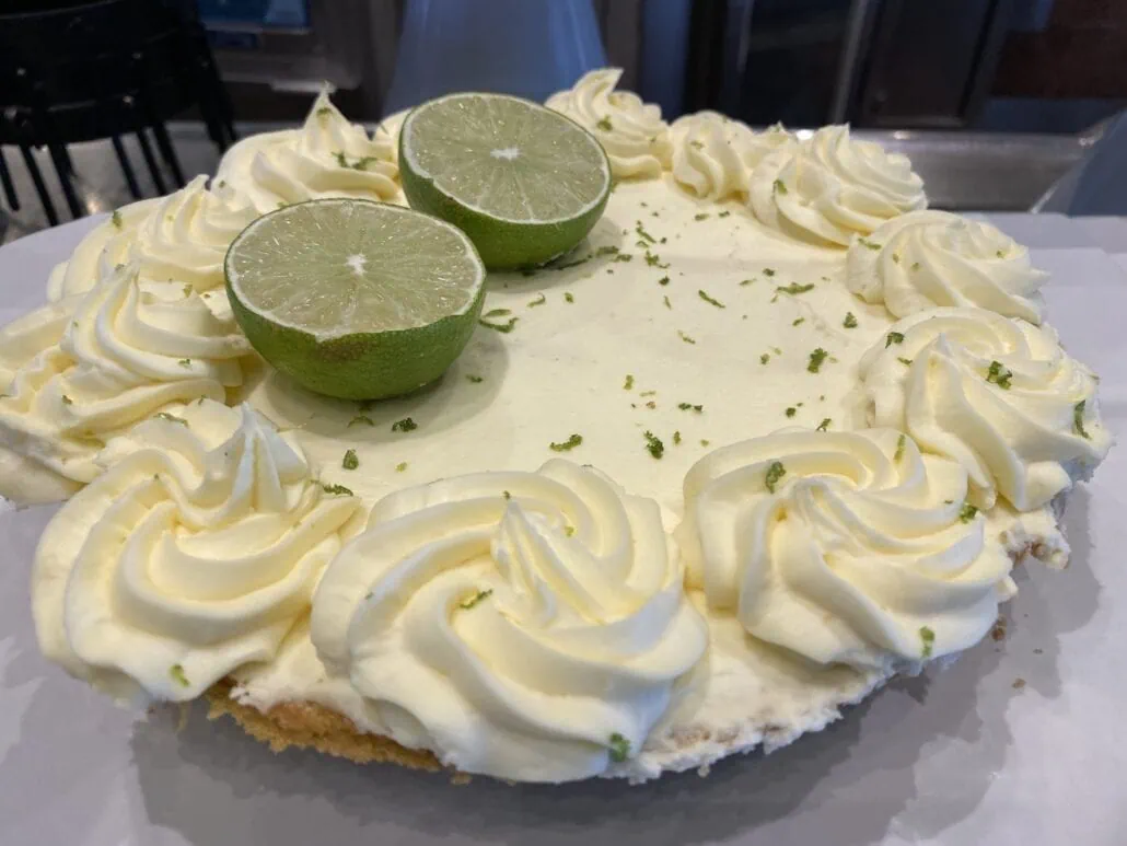 A key lime pie, a must-do activity for cruise passengers in Key West, is topped with white cream swirls around the edge and garnished with two halves of a fresh lime at the center. The pie sits enticingly on a pristine white surface.