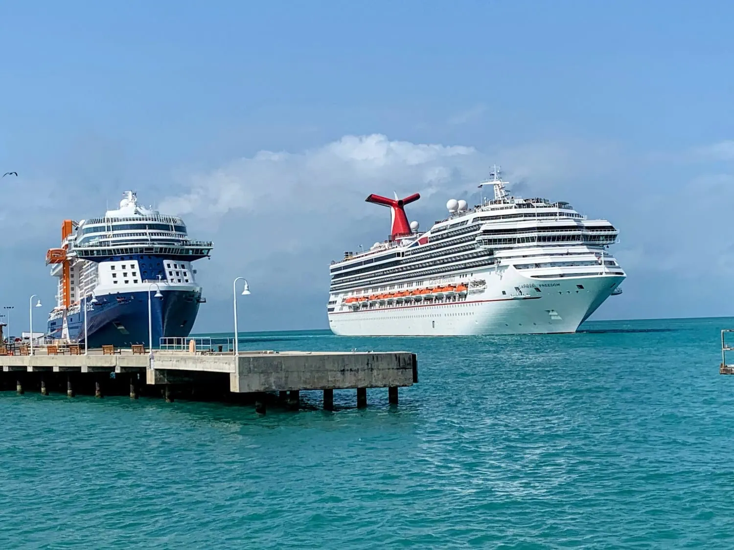 Two large cruise ships docked at a pier in the clear blue-green waters of Key West on a sunny day. The ship on the left is blue and white, while the ship on the right features a distinctive red and blue funnel. Cruise passengers enjoy various activities as they bask under a sky dotted with scattered clouds.