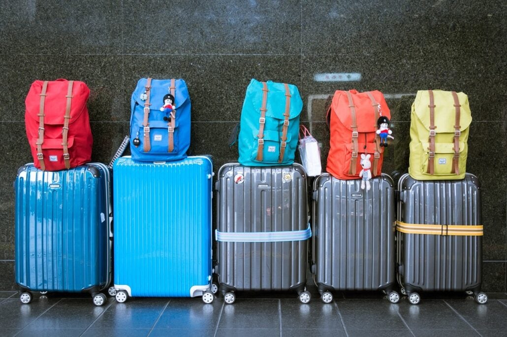 Five pieces of luggage with various colorful backpacks sit on top of them, arranged in a row against a dark wall. The backpacks are, from left to right, red, blue, turquoise, red, and yellow. Some bags have small toy decorations attached. A baggage handler might have some confessions about these unique items.