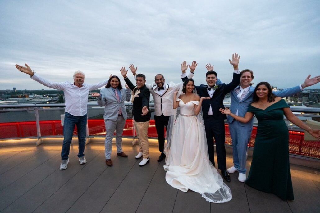 A group of eight people, including a bride and groom in wedding attire, stand on a rooftop terrace with arms raised, smiling and celebrating their surprise wedding aboard a Virgin Voyages ship.