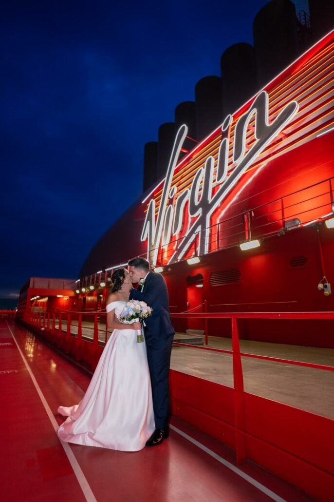 A couple in wedding attire embraces on a brightly lit Virgin Voyages cruise ship deck at night, capturing the essence of Sir Richard Branson's flair for surprise weddings.