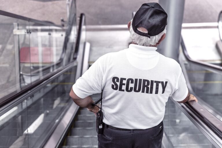 A security guard in a white shirt and black cap stands at the top of an escalator, overseeing the area, following a recent dispute over a cruise link drink package.
