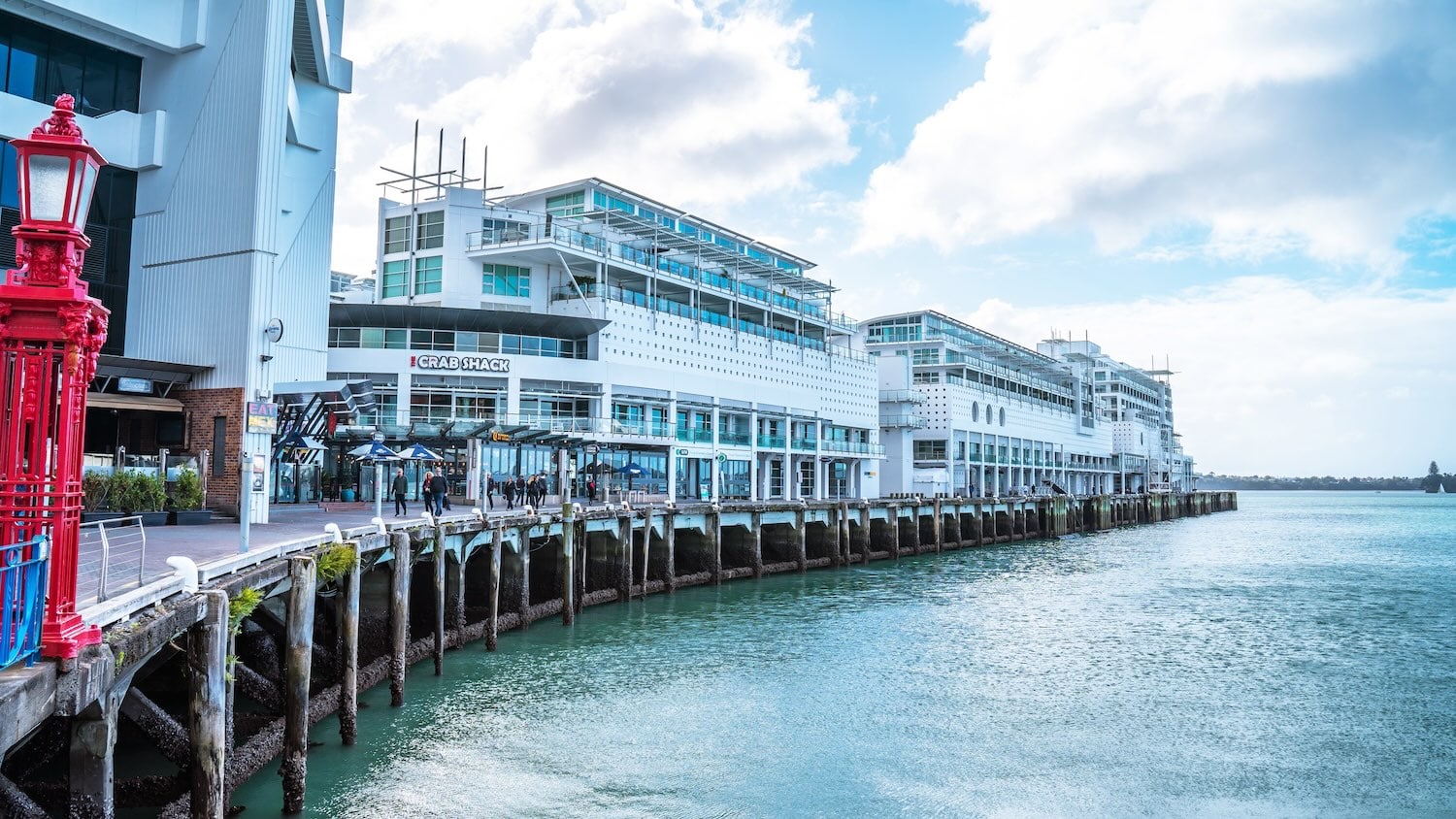 Image of a waterfront scene in New Zealand featuring modern white buildings with multiple floors, a restaurant named "1947 Eatery," and outdoor seating surrounded by a body of water under a partly cloudy sky.