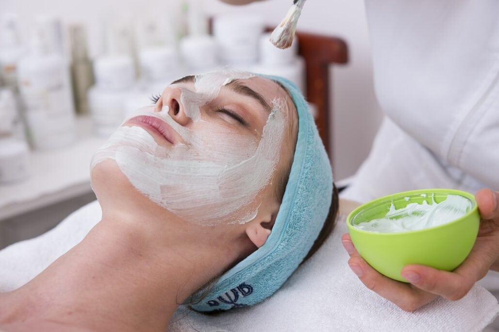 A person is lying back with their eyes closed, wearing a blue headband, while another applies a white facial mask with a brush. For travel tips on relaxing like this aboard cruise lines without breaking the bank, consider reading up on money-saving tips for your next journey.